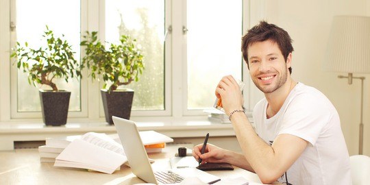 Confident smiling young man in white t-shirt sitting at his desk working on a laptop.