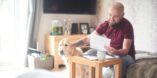 Leende man sitter hemma i soffan och håller ett papper i ena handen samtidigt som han klappar sin hund. 