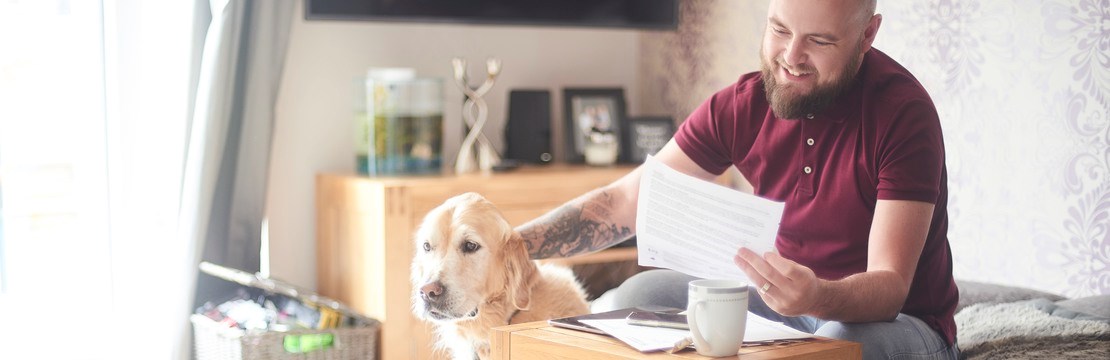 Leende man sitter hemma i soffan och håller ett papper i ena handen samtidigt som han klappar sin hund. 