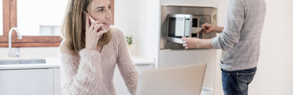 Kvinna jobbar och talar i mobil vid köksbordet  medan man värmer mat i mikrovågsugnen.
