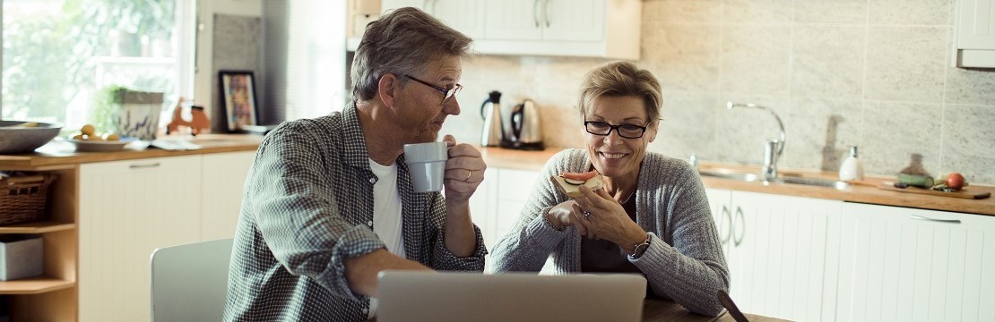 Ett par i övre medelåldern äter frukost i sitt kök med en laptop uppslagen på bordet.